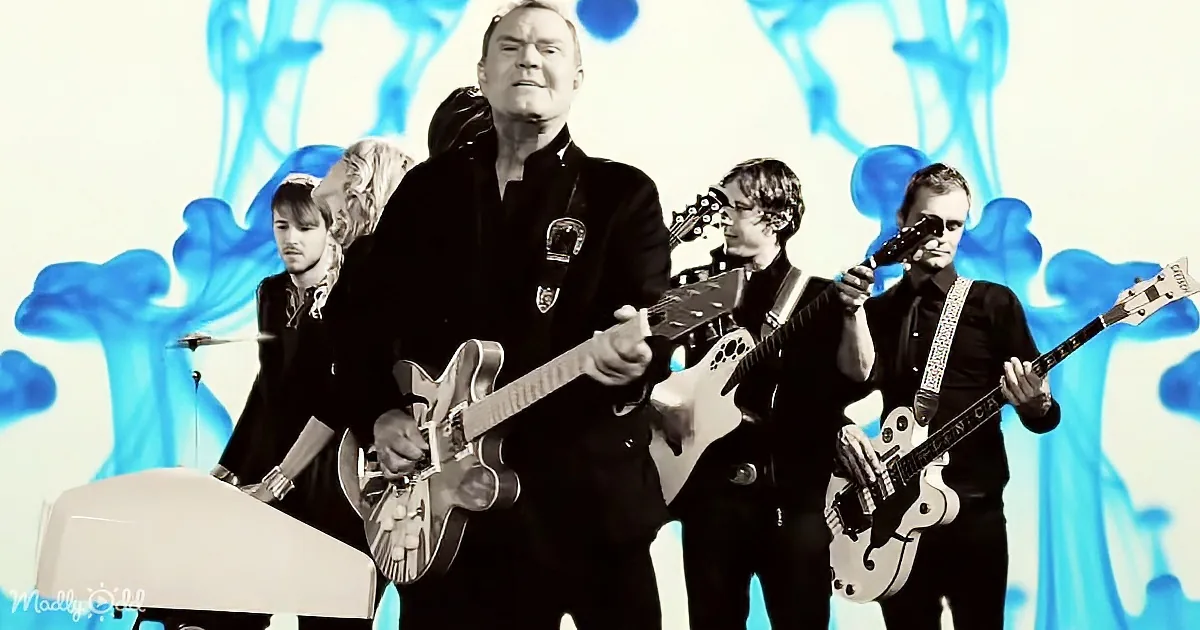 A man in a black suit plays a guitar on stage with a band, set against a blue abstract background.