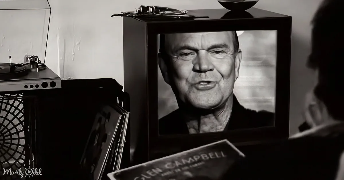A person holds a Glen Campbell album while a TV displays his face, with a record player and vinyl records in the background.
