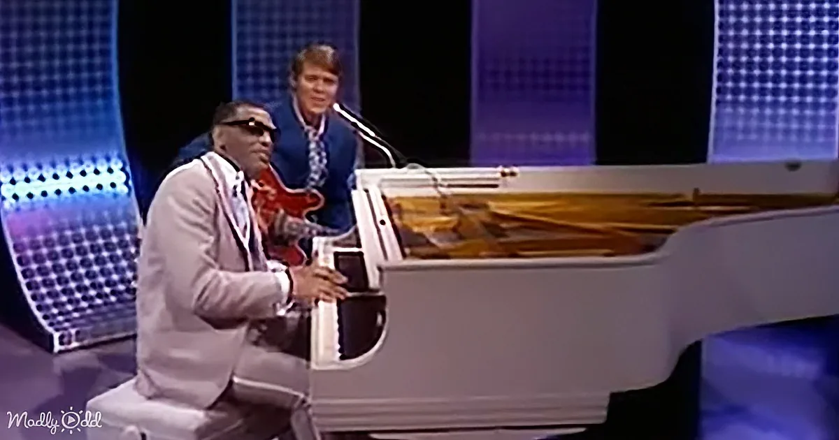 A man in a suit plays a white piano on a stage with blue lighting.