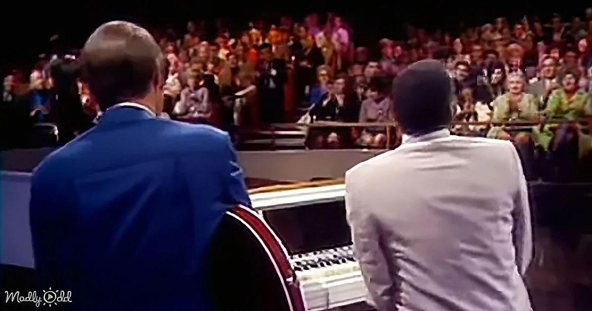 Two men in suits play a piano in front of a large audience.
