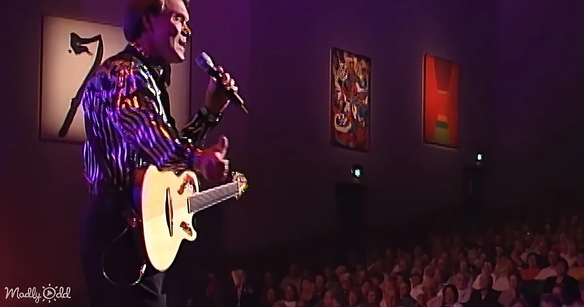 A man in a colorful shirt plays an acoustic guitar on stage, singing into a microphone.