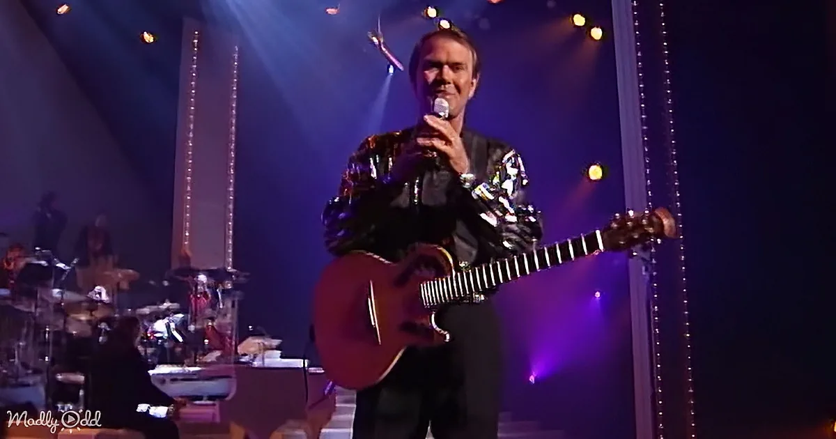 A man in a shiny jacket holds a microphone and guitar on stage with a band and colorful lights.