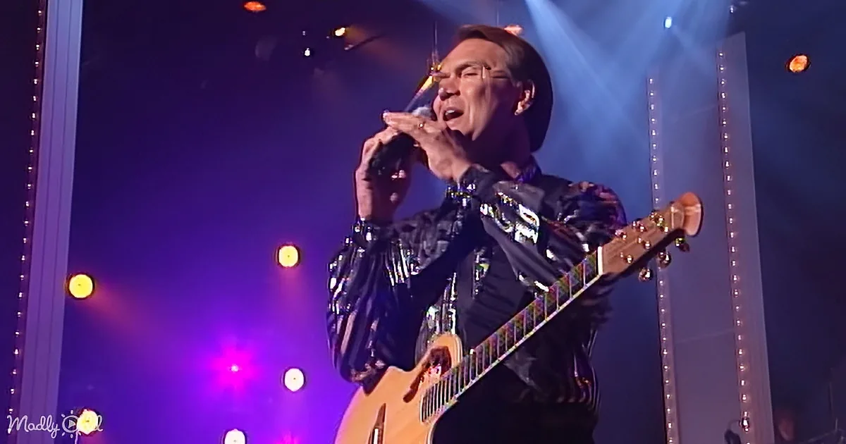 A person in a shiny outfit plays a guitar and sings on stage with colorful lights.