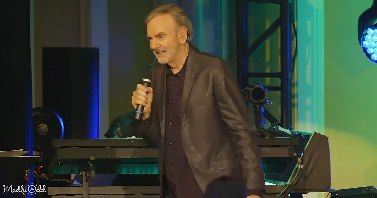 A man in a dark suit holds a microphone on stage, smiling, with a keyboard and stage lights in the background.