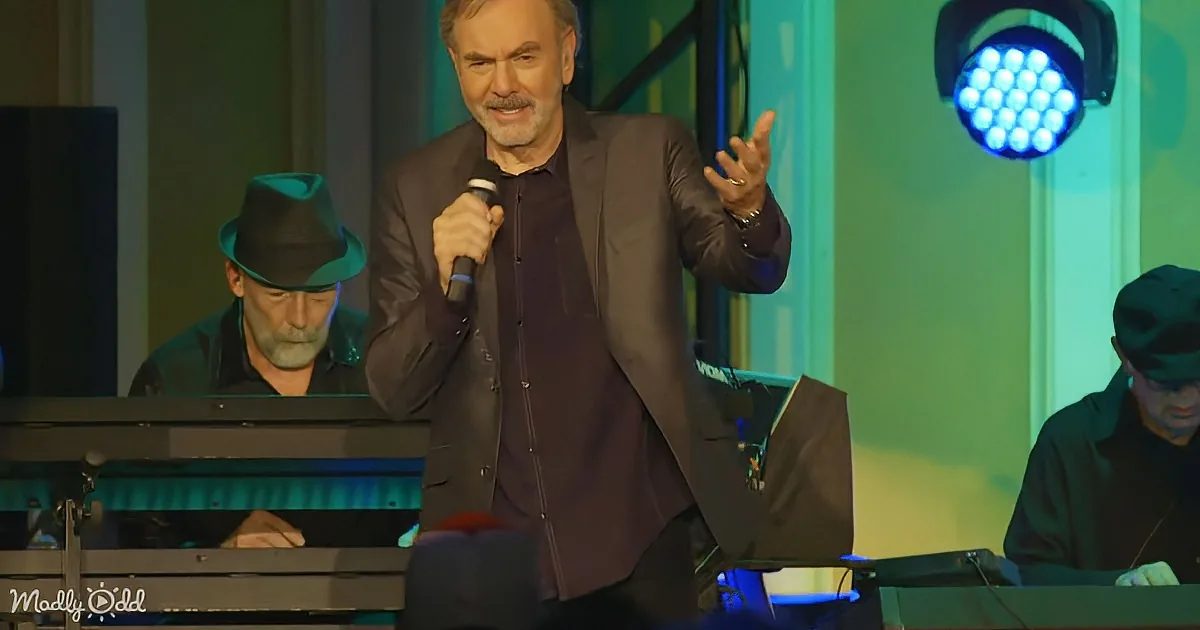 A man in a suit holds a microphone, gesturing with his other hand, while musicians play keyboards in the background.