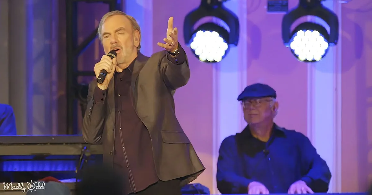 A man in a suit sings into a microphone, gesturing with his hand, while another man plays a keyboard in the background.