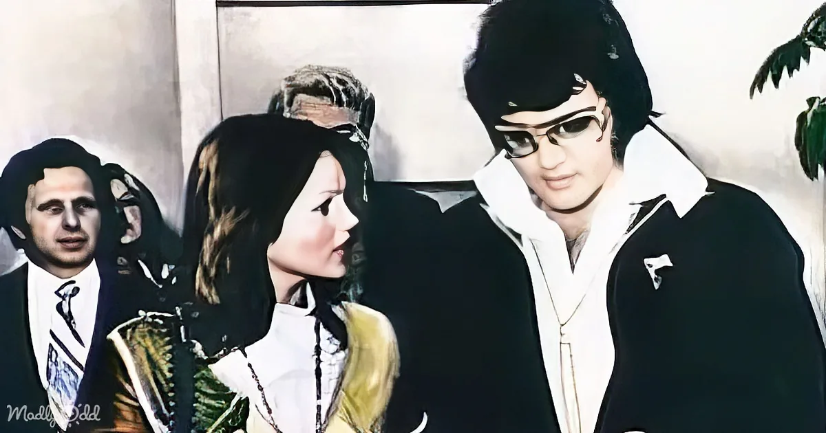 Elvis Presley in a black and white outfit with sunglasses, standing next to a woman in a white and gold dress.