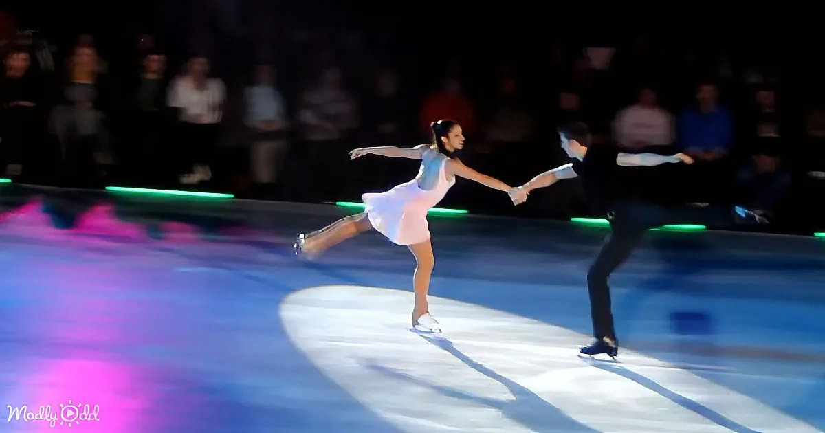 A figure skater in a white dress glides gracefully on ice, holding hands with a partner.