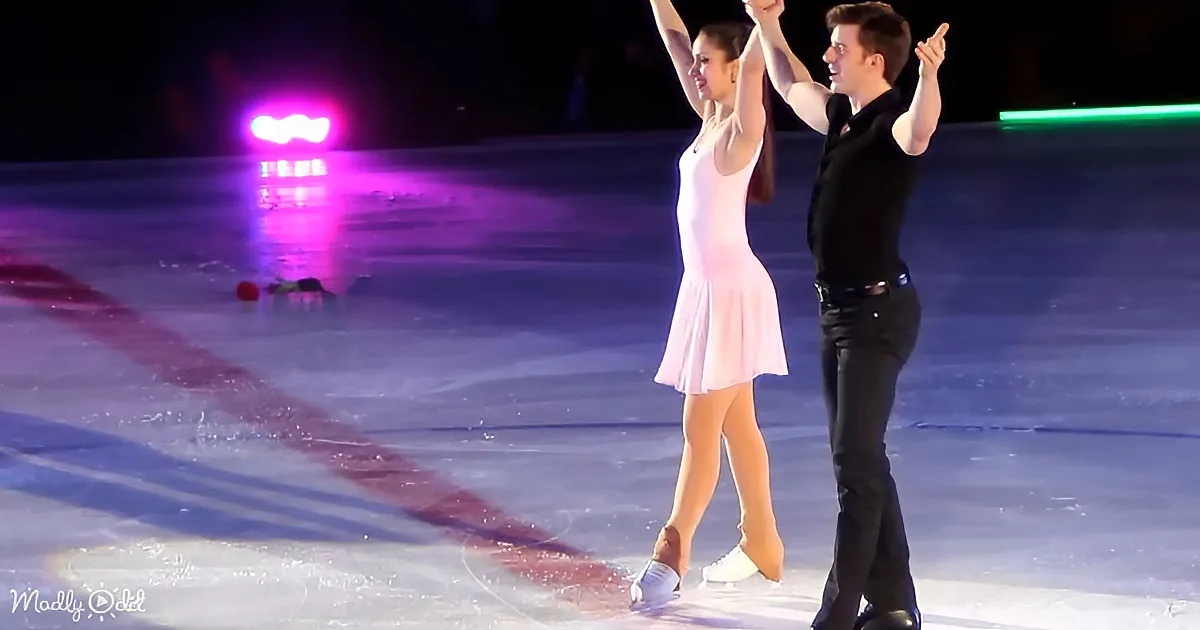 A figure skating couple in pink and black holds hands, gliding on ice with colorful lights in the background.