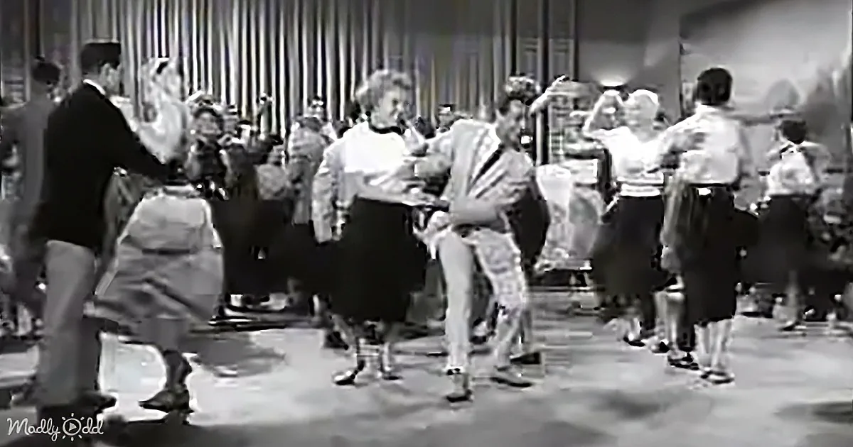 A man in a suit dances with a woman in a skirt, surrounded by other couples in a lively dance scene.