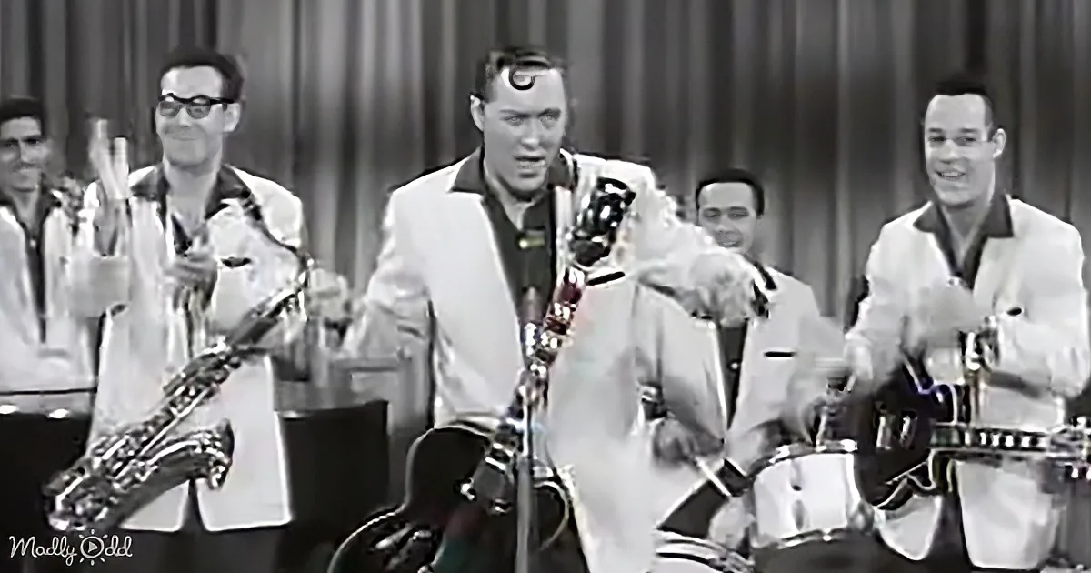 A man in a white suit plays guitar on stage with a band, including saxophonists and a guitarist.
