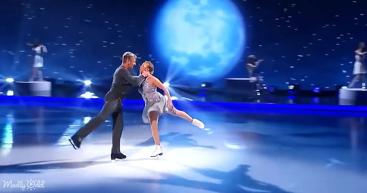 A couple in a graceful ice skating pose, illuminated by stage lights, with a large moon backdrop.