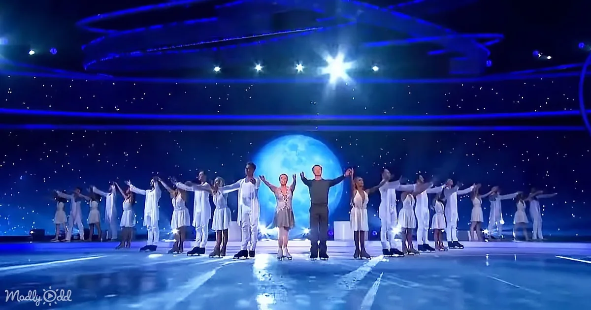 A group of performers in white costumes skate on ice, arms outstretched, under a starry sky backdrop.