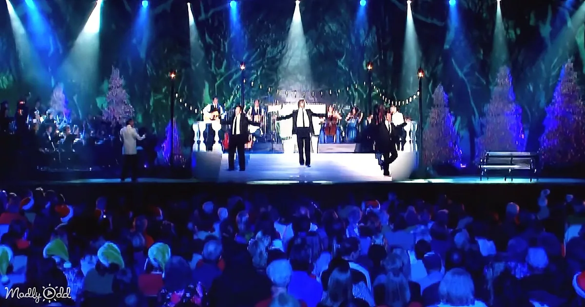 A stage with performers and orchestra under blue lights, surrounded by a festive audience.