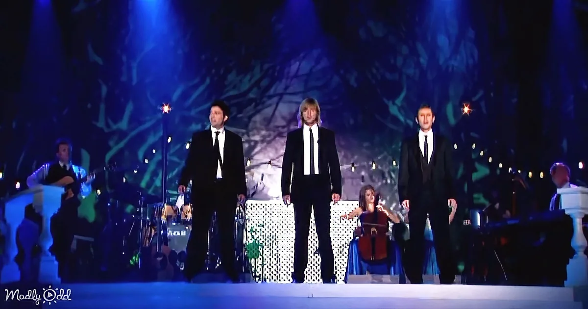 Three men in black suits stand on stage, illuminated by blue lights, with musicians in the background.
