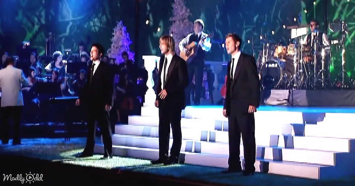 Three men in suits sing on stage with a band and orchestra in the background.