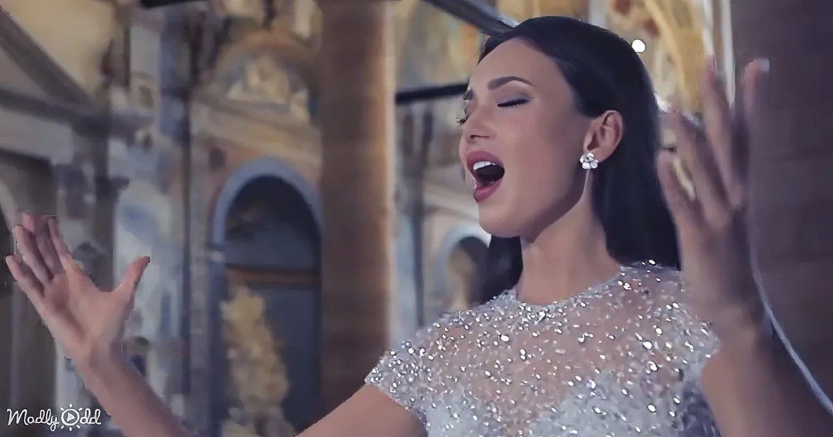 A woman in a sparkling dress sings passionately with her hands raised, set against a blurred architectural background.