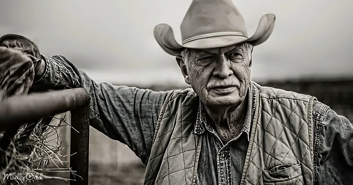 An older man in a cowboy hat and quilted vest leans on a fence, gazing into the distance.