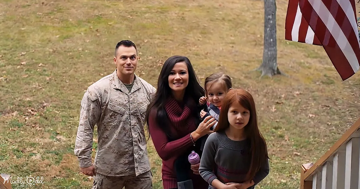 A family of four stands outdoors, with a man in military uniform, a woman holding a child, and another child standing nearby.