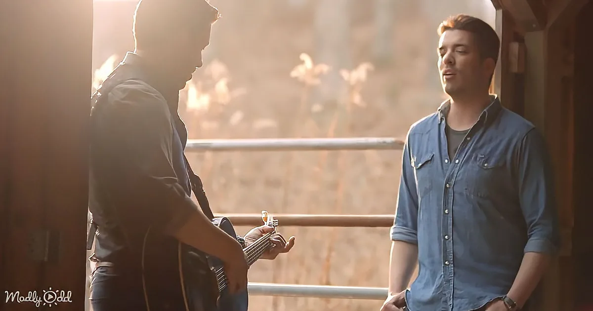 A man in a denim shirt stands near a railing, looking at another man playing a guitar.