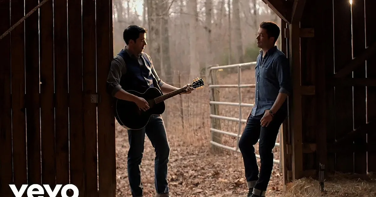 A man plays guitar while another listens, both in a rustic setting with wooden beams and a railing.
