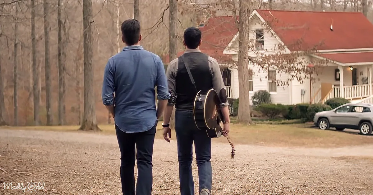 Two men walk away, one carrying a guitar, towards a house with a red roof and bare trees.