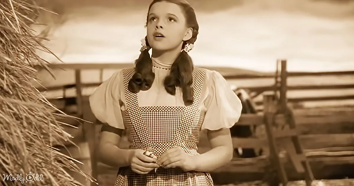 A woman in a checkered dress stands outdoors, looking upward, with a haystack and wooden structure in the background.