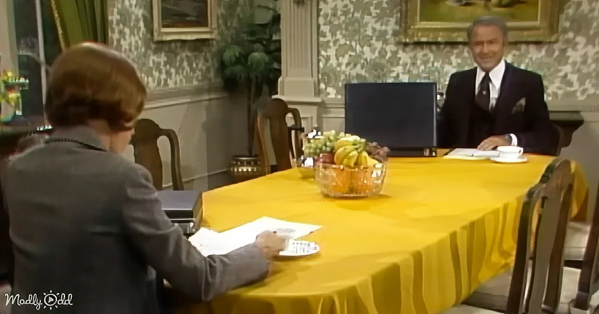 A man in a suit sits at a table with a yellow tablecloth, smiling, while another person in a suit works at a desk nearby.