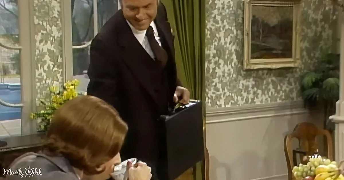 A man in a suit smiles while holding a briefcase, standing near a seated person in a cozy room with floral wallpaper.