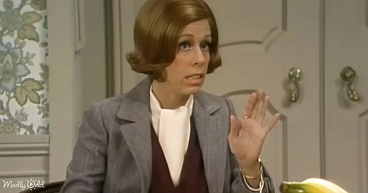 A woman in a gray blazer and maroon sweater gestures with her right hand, appearing to speak or explain something.