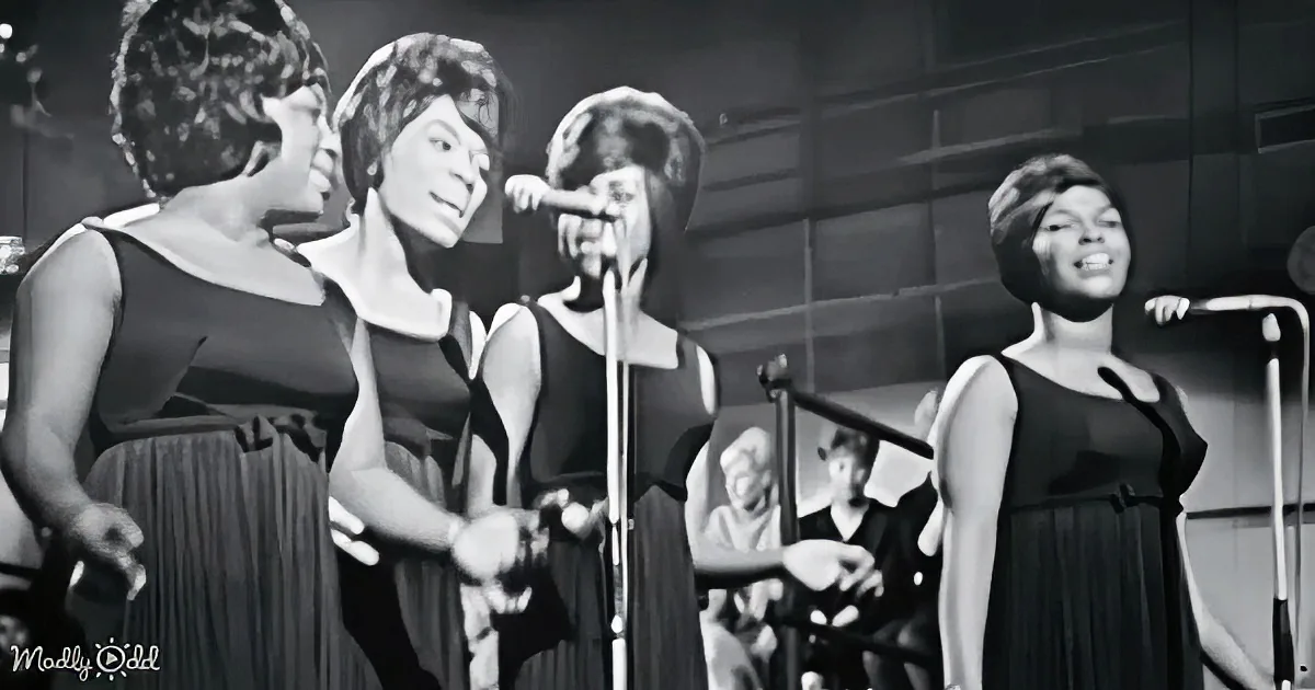 Four women in black dresses sing on stage, one holding a microphone, with a band in the background.