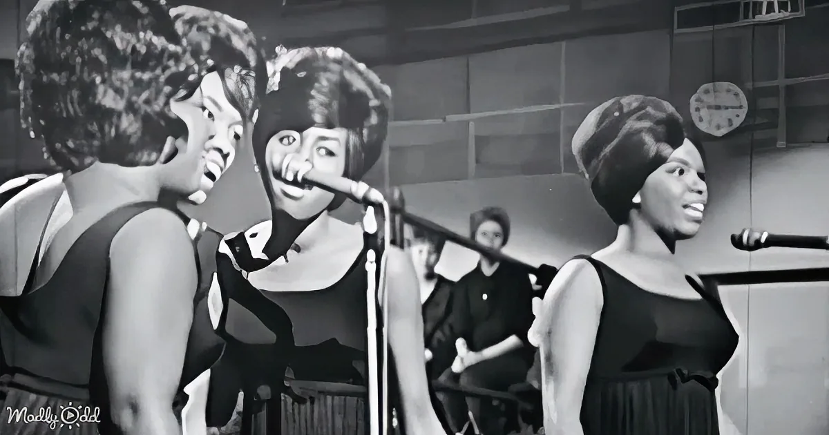 A woman in a black dress sings into a microphone, accompanied by two other women in similar attire.