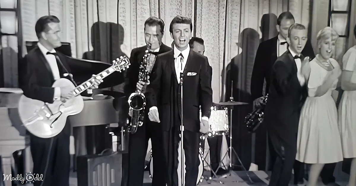 A man in a suit sings into a microphone, surrounded by musicians and a choir in formal attire.