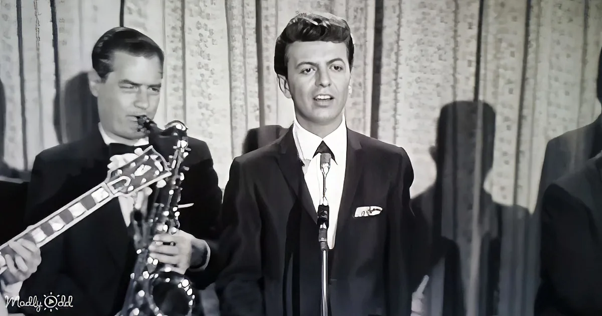 A man in a suit sings into a microphone while another plays a saxophone in the background.