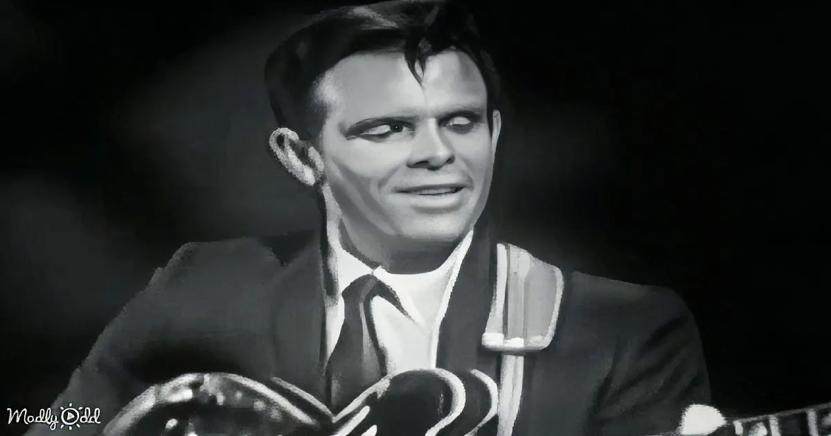 A man in a suit and tie is speaking into a microphone, with a dark background.