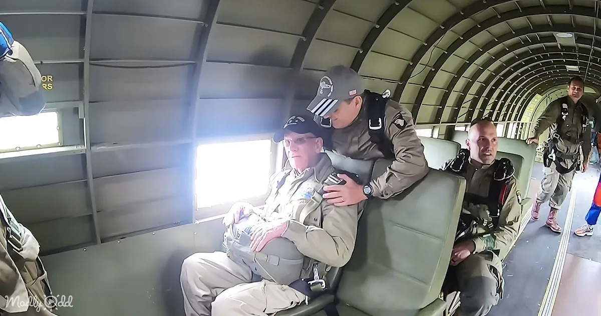 A man in a military uniform sits in a plane, being supported by another man in a similar uniform.