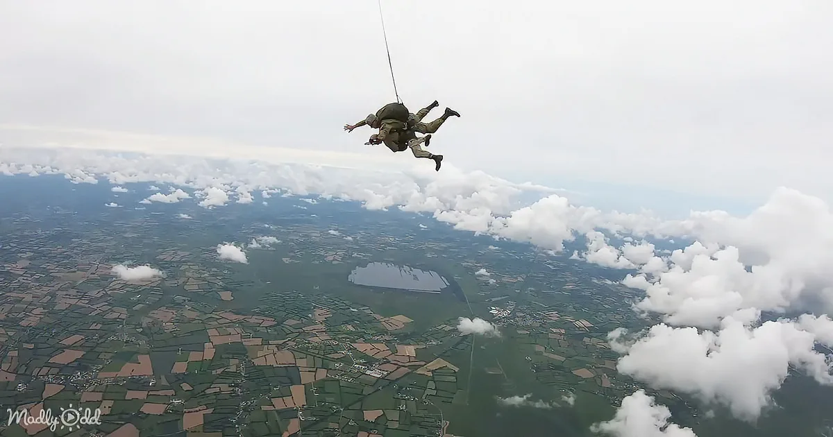 A skydiver descends with a parachute, suspended above a patchwork landscape of fields and clouds.