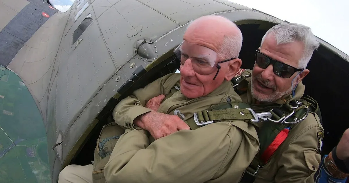 Two men in green flight suits are seated inside a cockpit, one holding the edge of the open door.
