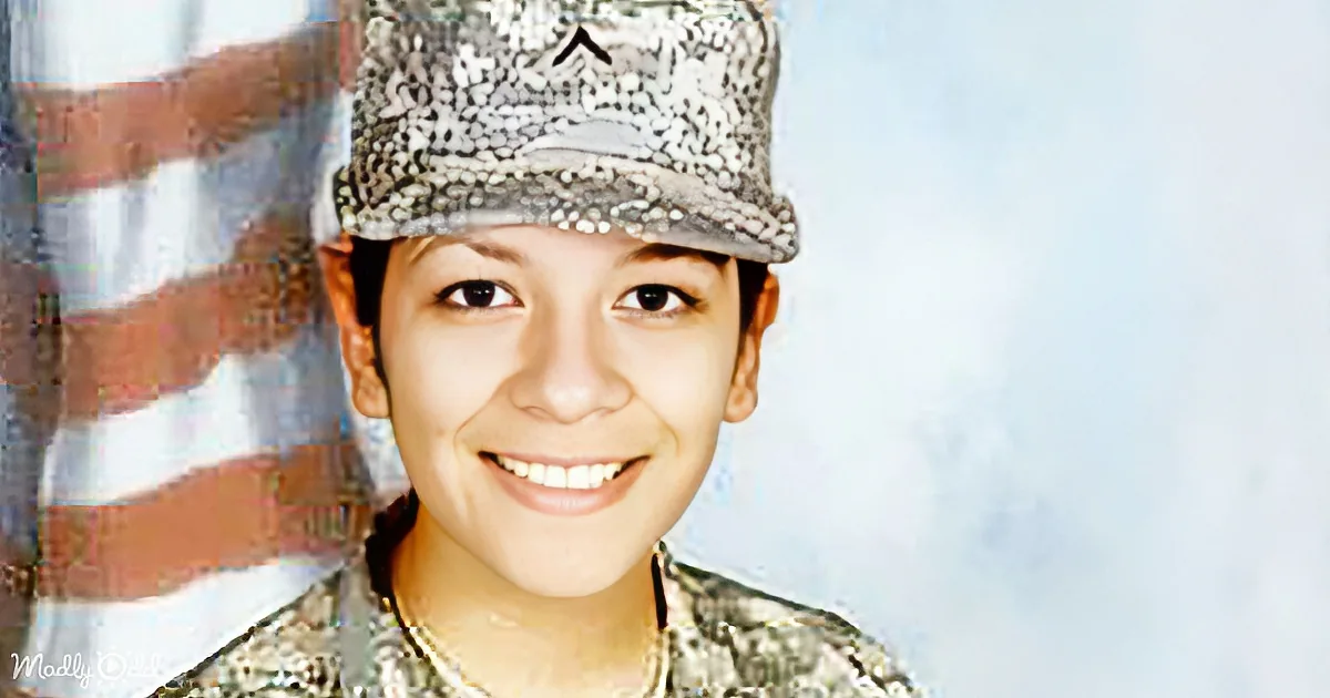 A smiling person in military uniform with a cap, standing near an American flag.