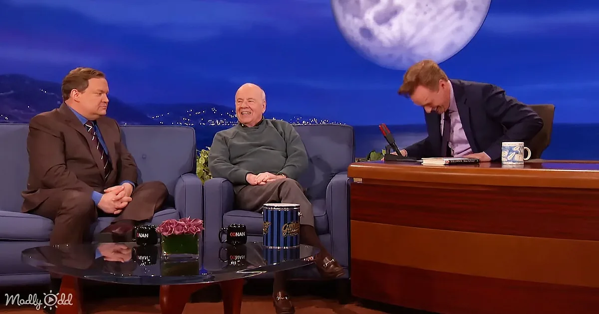 Conan O'Brien sits at desk, interviewing two guests on a talk show set with a large moon backdrop.