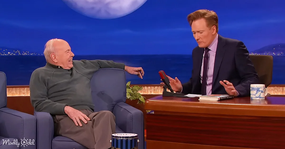 A man in a green sweater sits relaxed in a blue chair, laughing, while another man in a suit gestures during a talk show.