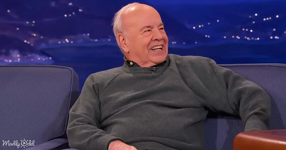 An older man in a dark green sweater sits on a blue couch, smiling with a starry background.