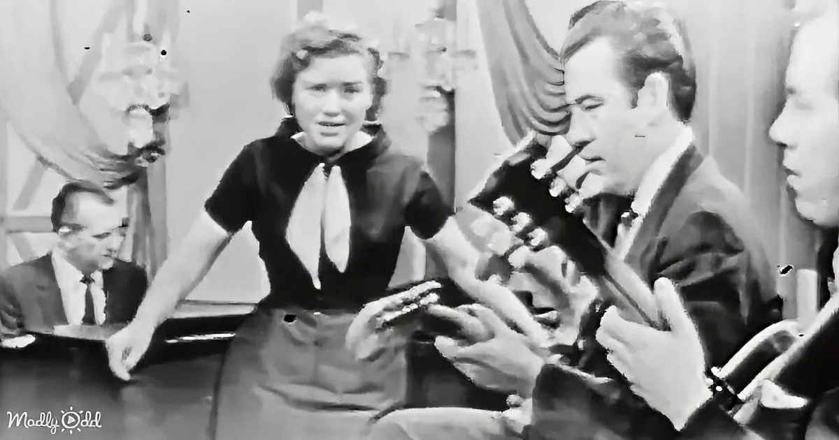 A woman in a black dress and white bow tie is holding a guitar, surrounded by people in a formal setting.