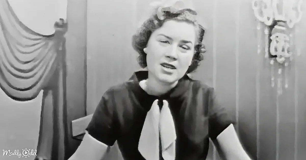 A woman in a black outfit with a white bow tie stands in a room with a draped curtain and decorative wall.