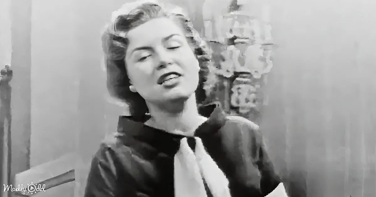 A woman with curly hair, wearing a dark outfit, stands indoors, possibly singing or speaking.