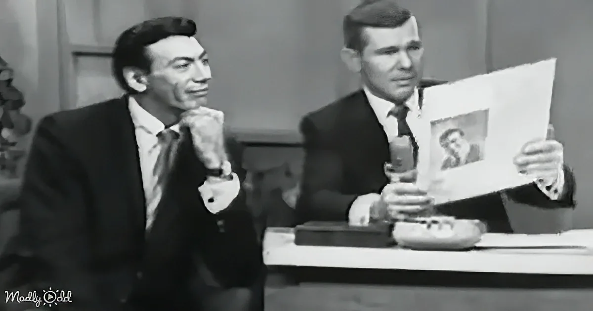 A man in a suit holds a photo while another man in a suit leans on a table, appearing thoughtful.