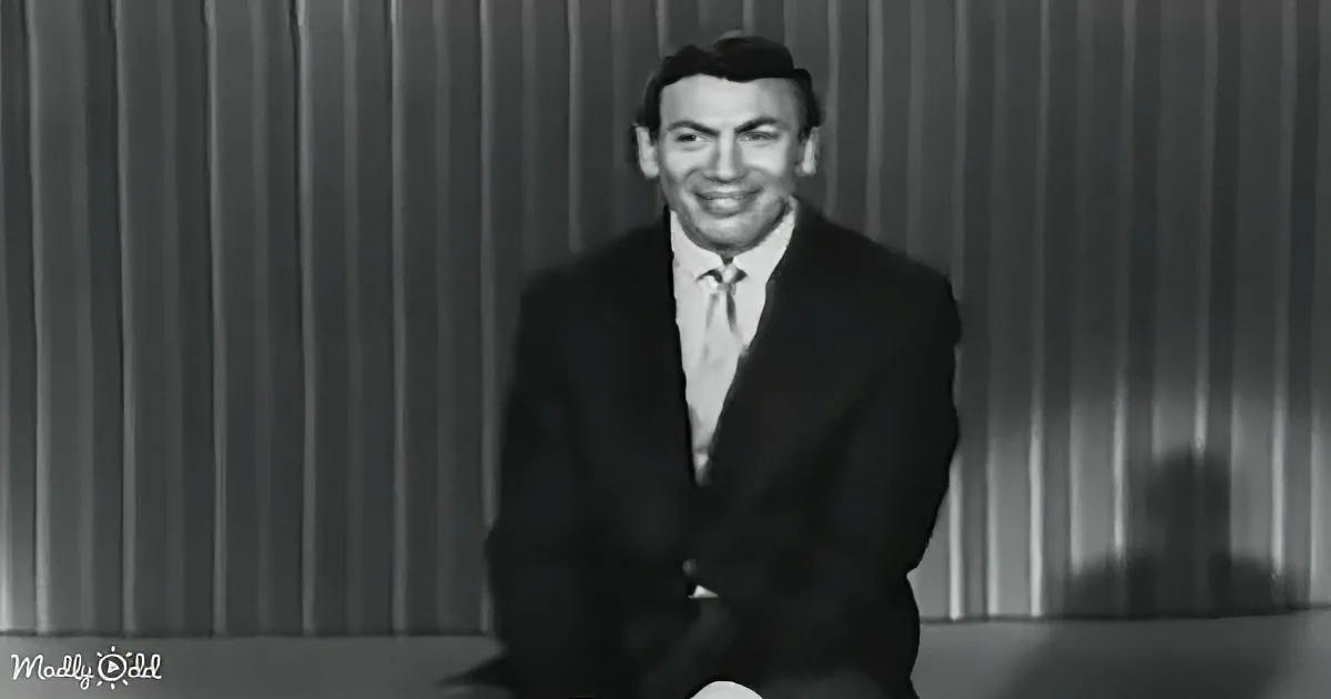 A man in a suit and tie sits on a stage, smiling, with a striped curtain backdrop.