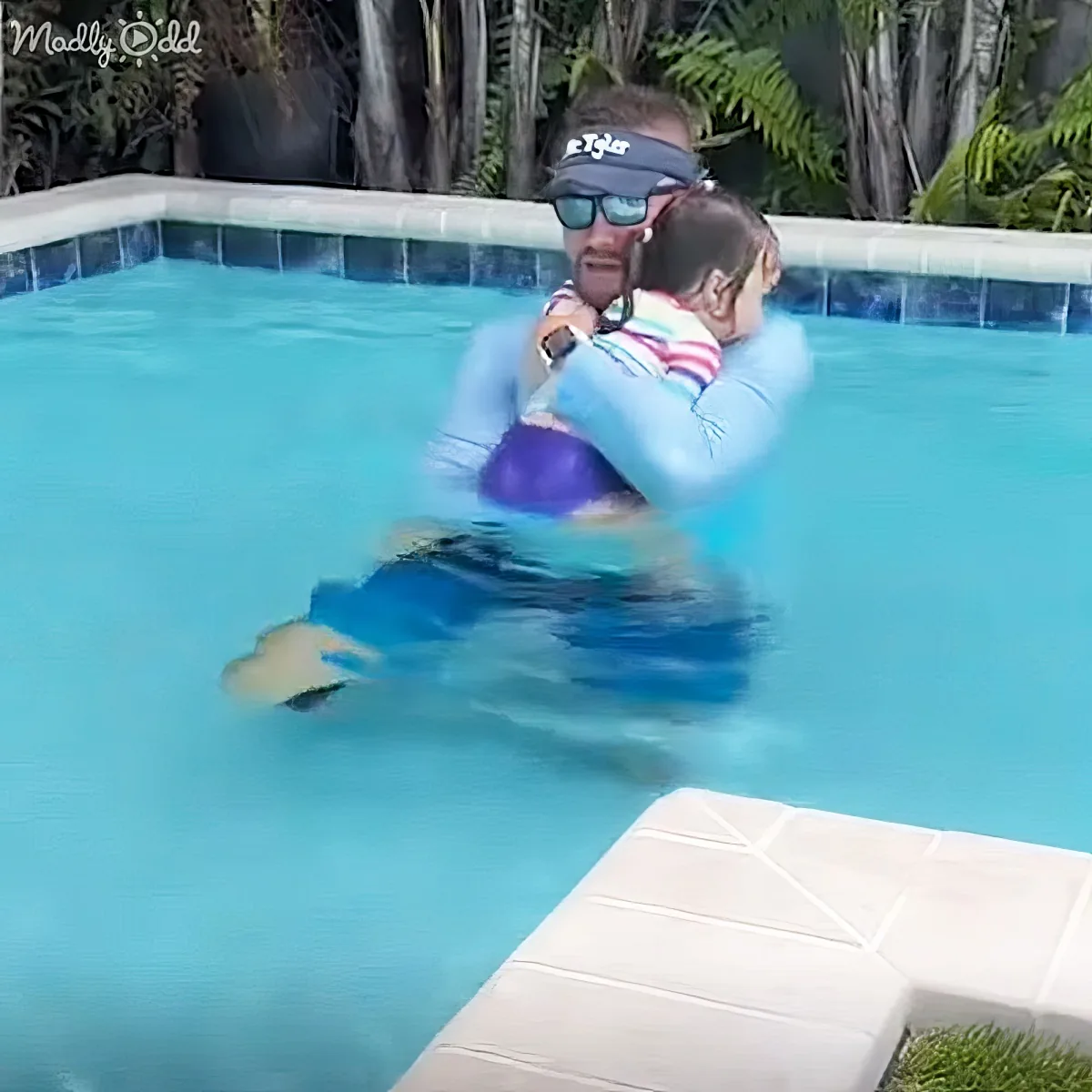 A man holds a child in a pool, both wearing swim gear, with a tropical backdrop.