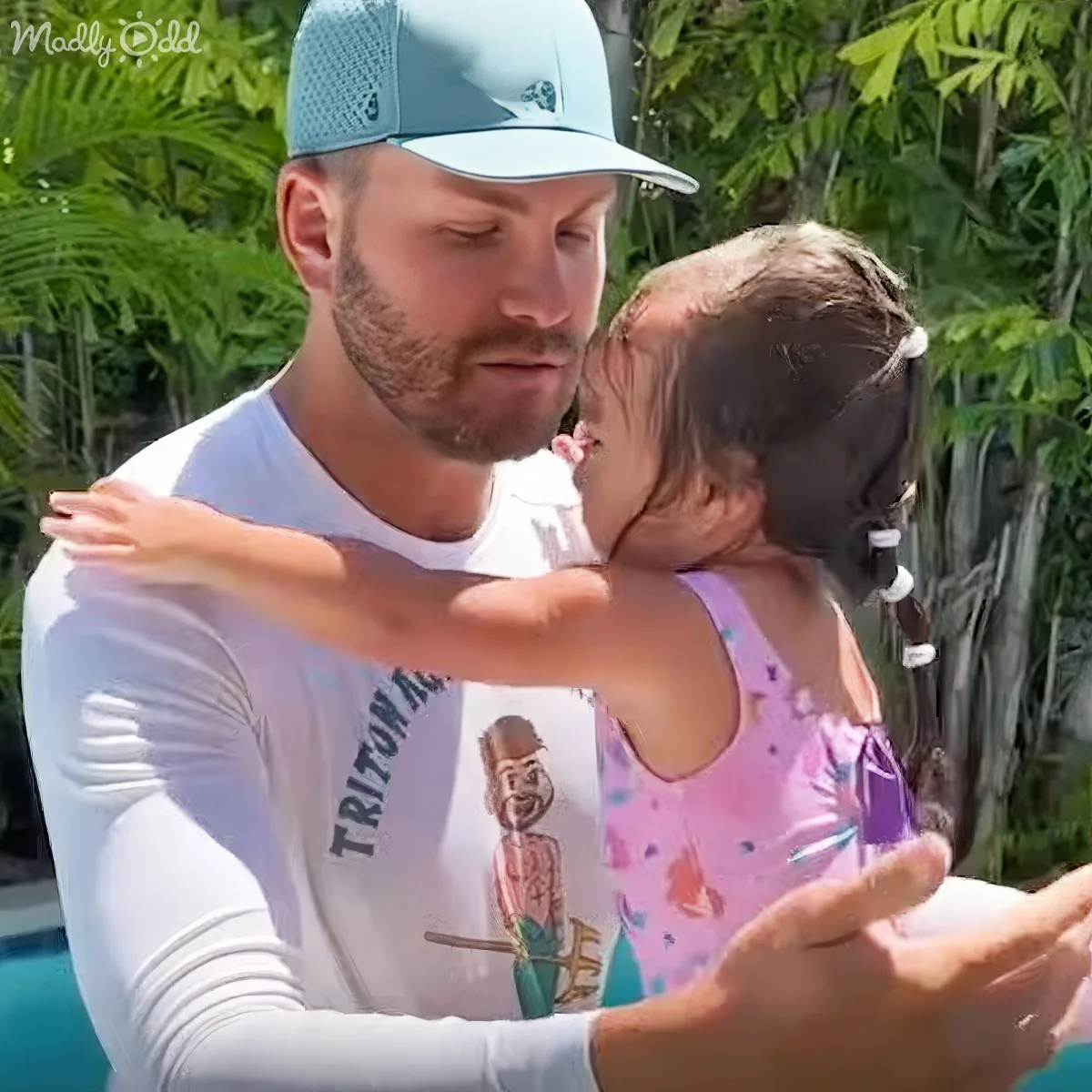 A man in a white shirt and blue cap holds a child in a pink swimsuit, both outdoors with greenery in the background.