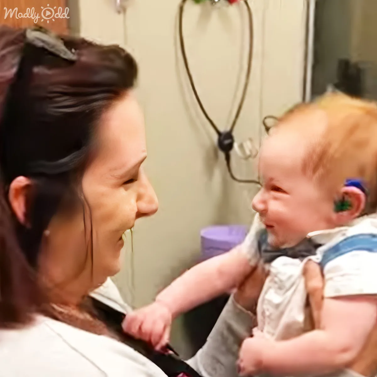 A baby with a hearing aid smiles while being held by a woman in a clinical setting.